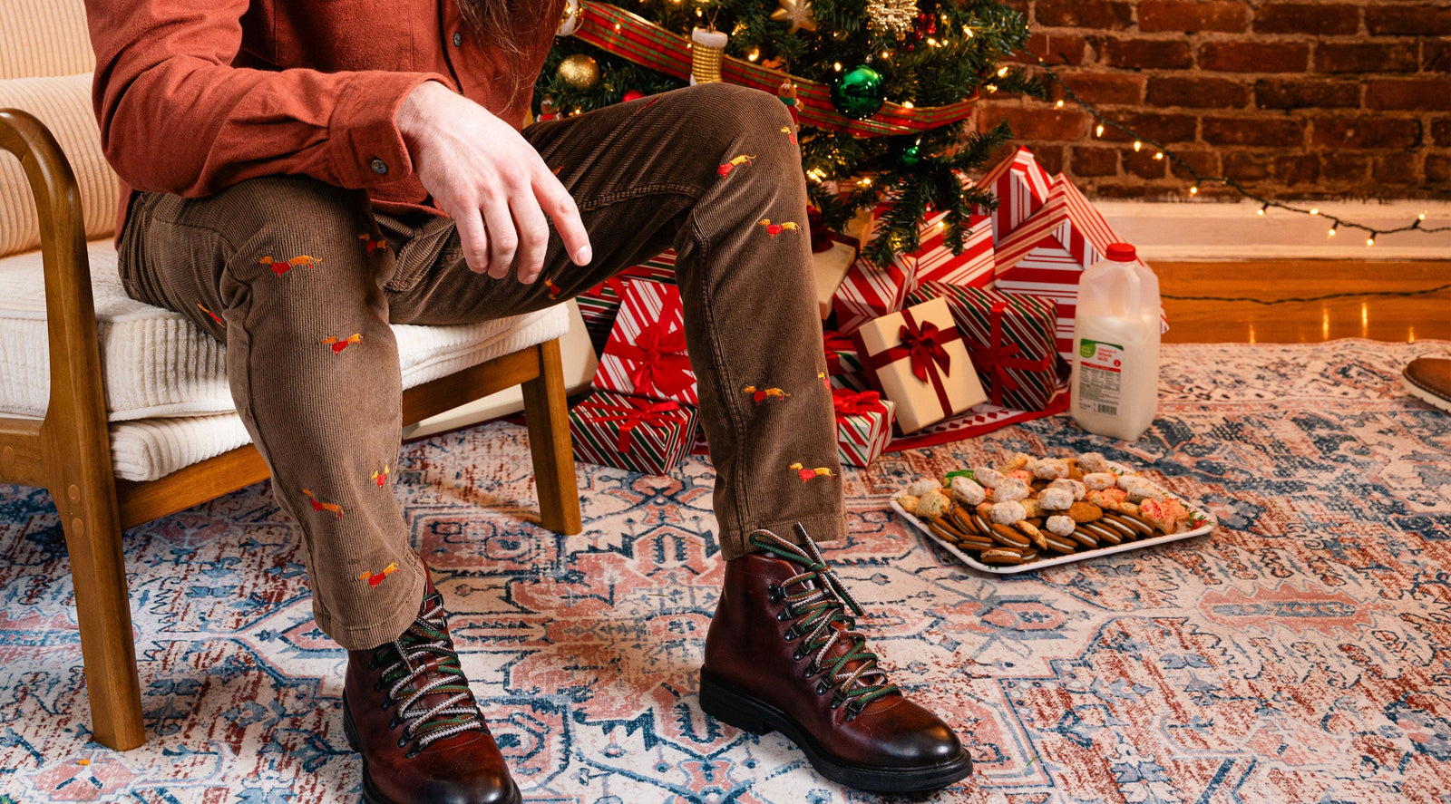 Person wearing brown corduroy pants embroidered with dachshunds and brown boots sitting near a Christmas tree with wrapped gifts, a plate of cookies, and a milk jug on a patterned rug.