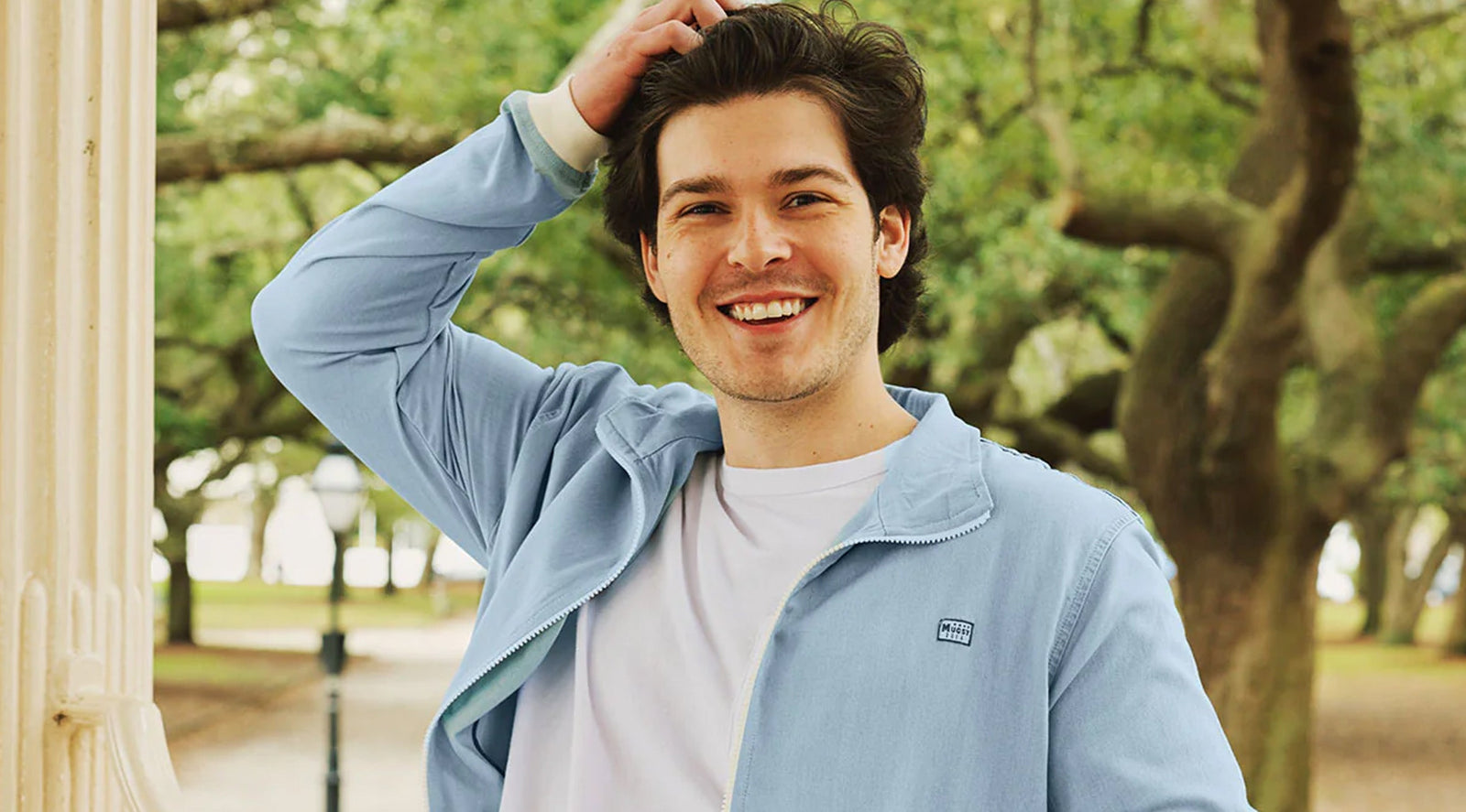 Smiling man outdoors wearing a light blue jacket with a Mugsy logo patch on the chest, standing near a column with trees in the background