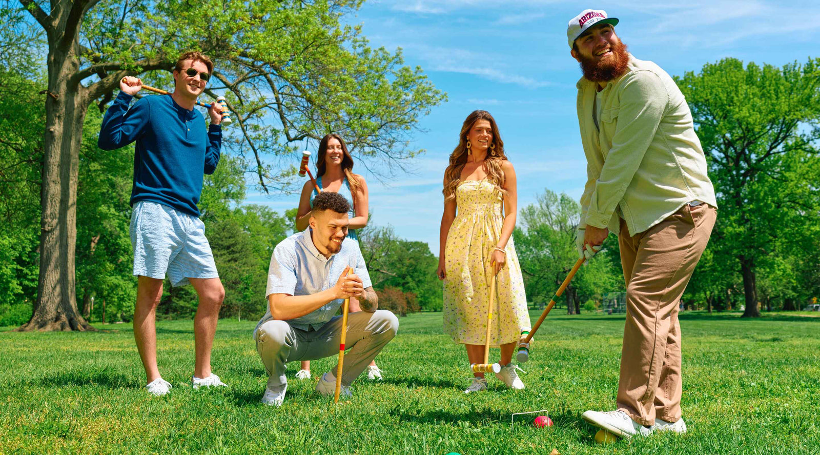 Five people playing croquet on a sunny day in a park with green trees and grass