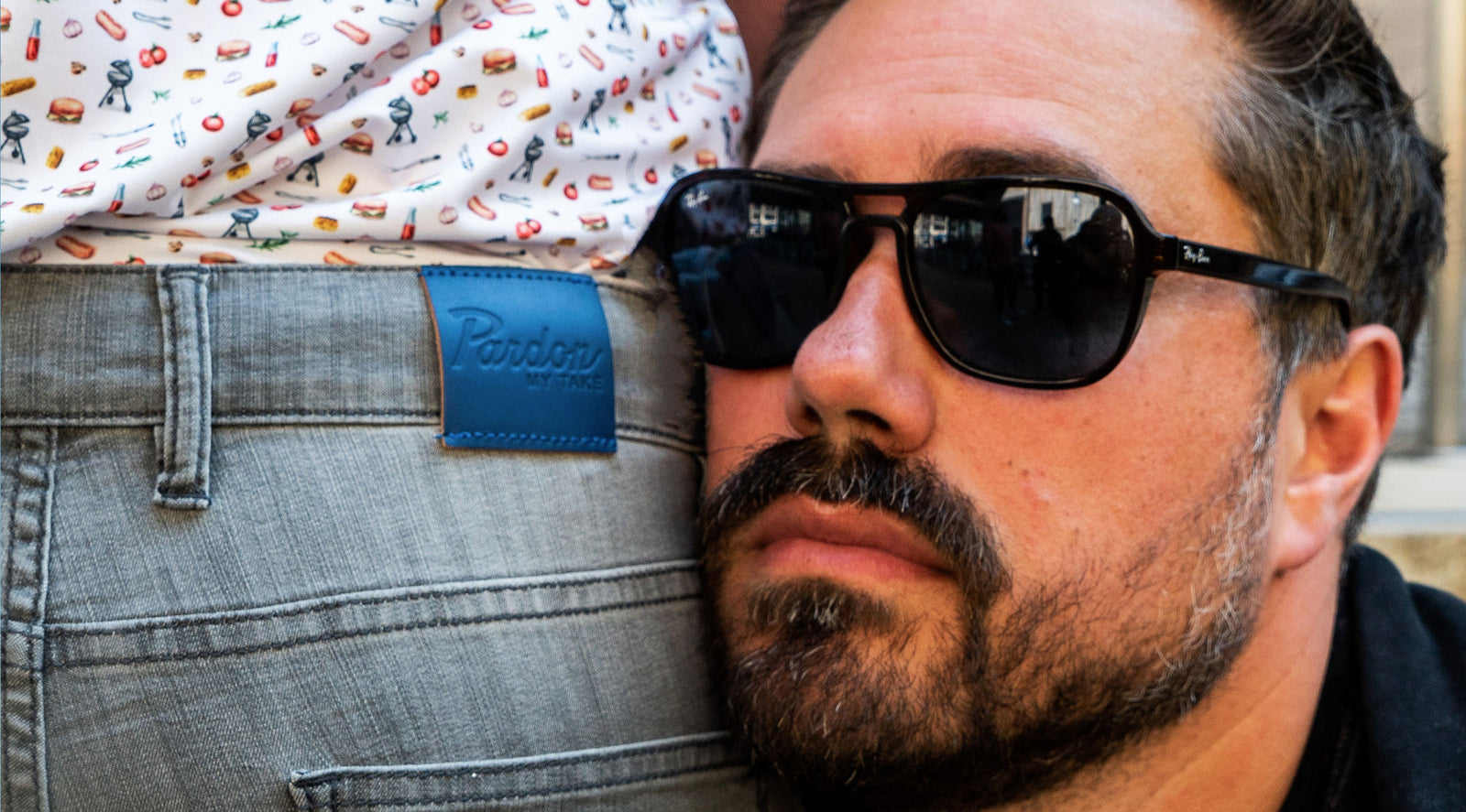 Man wearing black sunglasses next to a person in gray jeans with a blue label reading 'Pardon MY TAKE'
