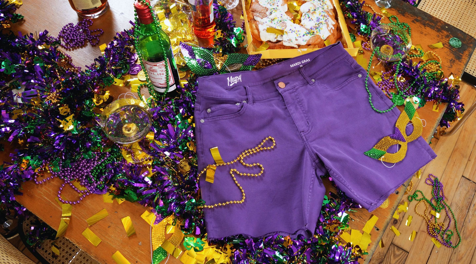 Top-down view of purple Mugsy cut-off shorts showing raw frayed hem and five-pocket styling on Mardi Gras beads and confetti