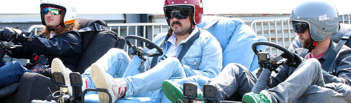 Three men wearing helmets and sunglasses sitting in go-karts, each with distinct colored helmets and casual clothing, preparing for a race or ride.