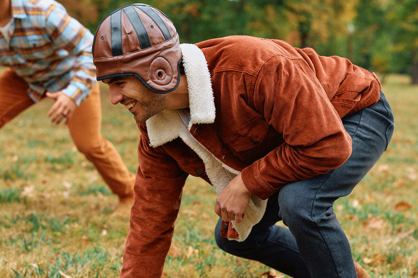 Man wearing a brown corduroy jacket with white fleece lining and a brown leather helmet crouching on grass outdoors, with another person blurred in the background.