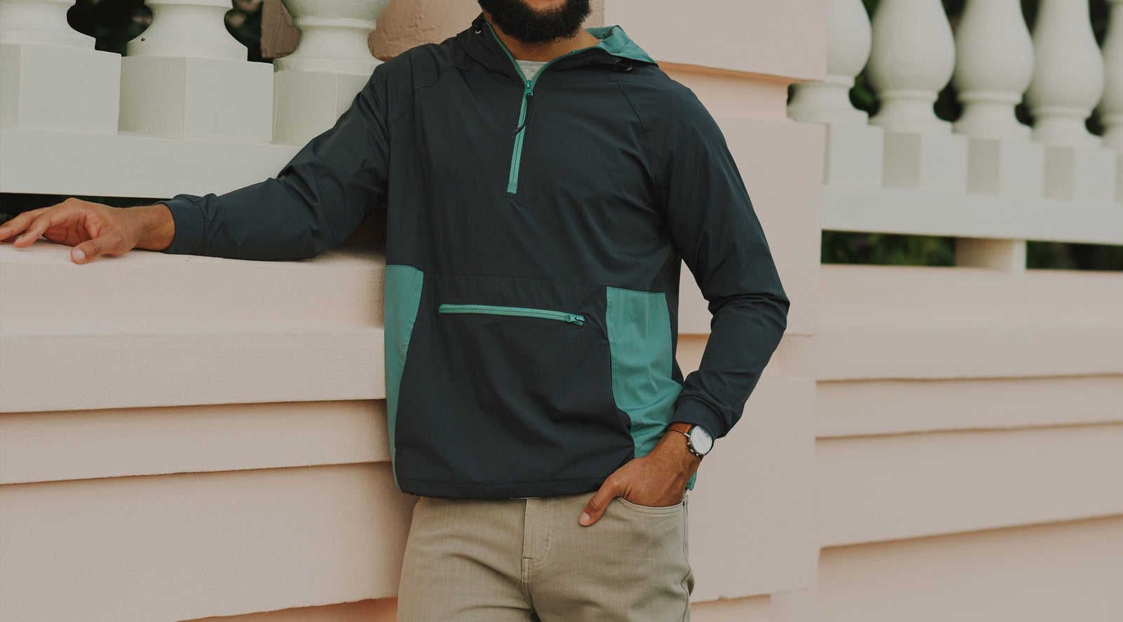 Man wearing a black and teal hooded jacket with a front zipper pocket, standing against a beige wall with white balusters