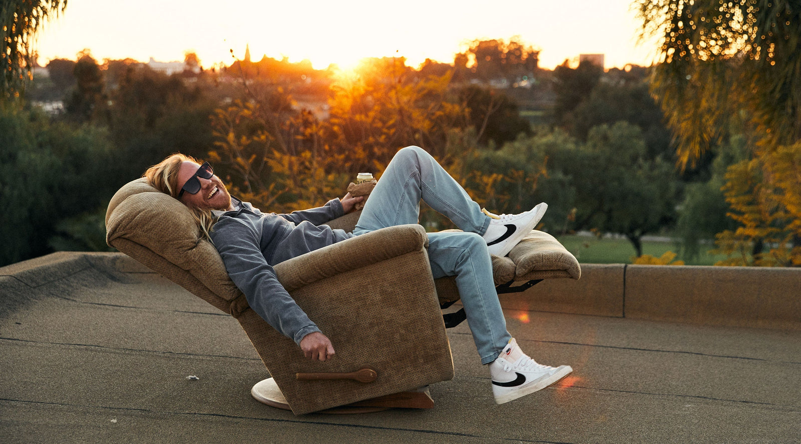 Man reclining in a brown recliner chair outdoors at sunset, smiling and wearing sunglasses, gray sweatshirt, light blue jeans, and white Nike sneakers.