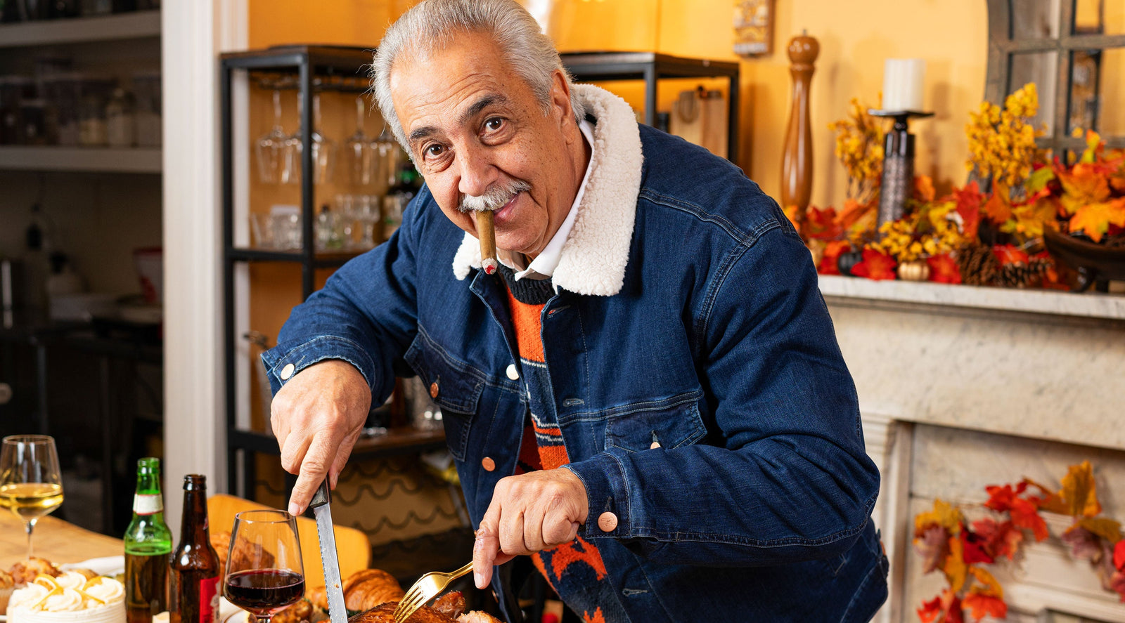 Older man with gray hair and mustache, wearing a denim jacket and orange sweater, carving meat at a festive table with autumn decorations and drinks, holding a cigar in his mouth.
