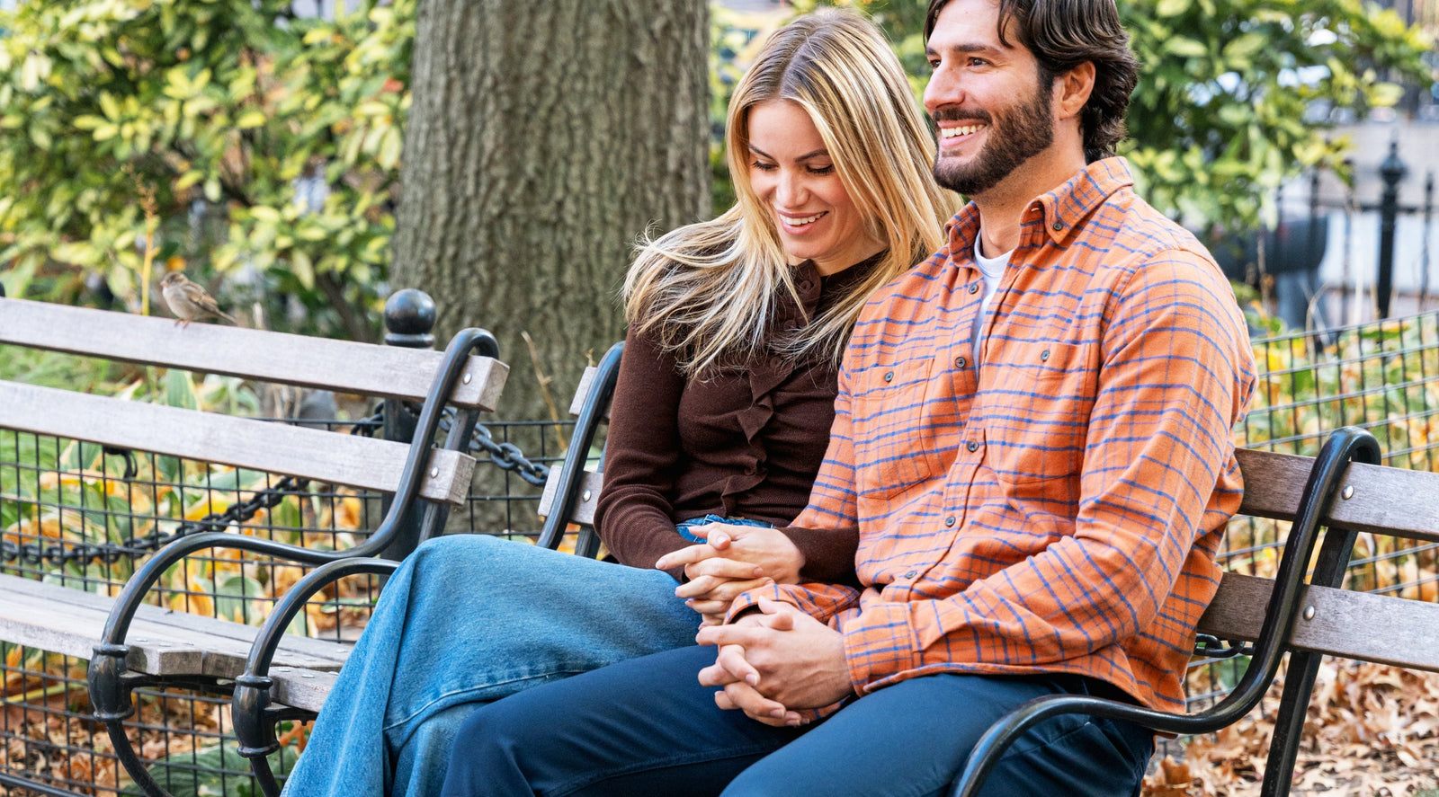 Smiling couple sitting on a park bench holding hands surrounded by fall foliage.