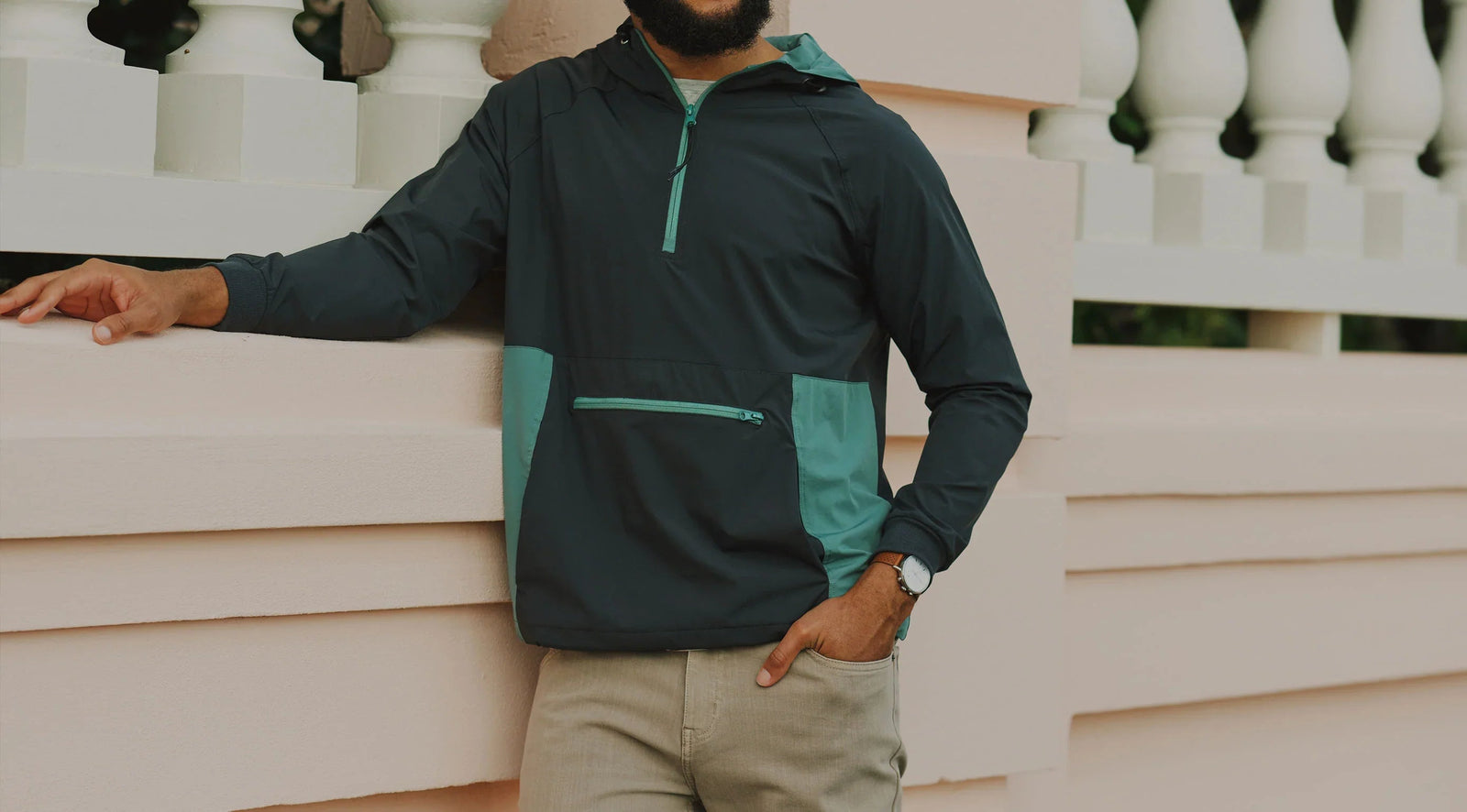Front view of a men's dark teal pullover windbreaker with mint-green accents, half-zip hood and zippered kangaroo pocket, model leaning on pale pink balustrade
