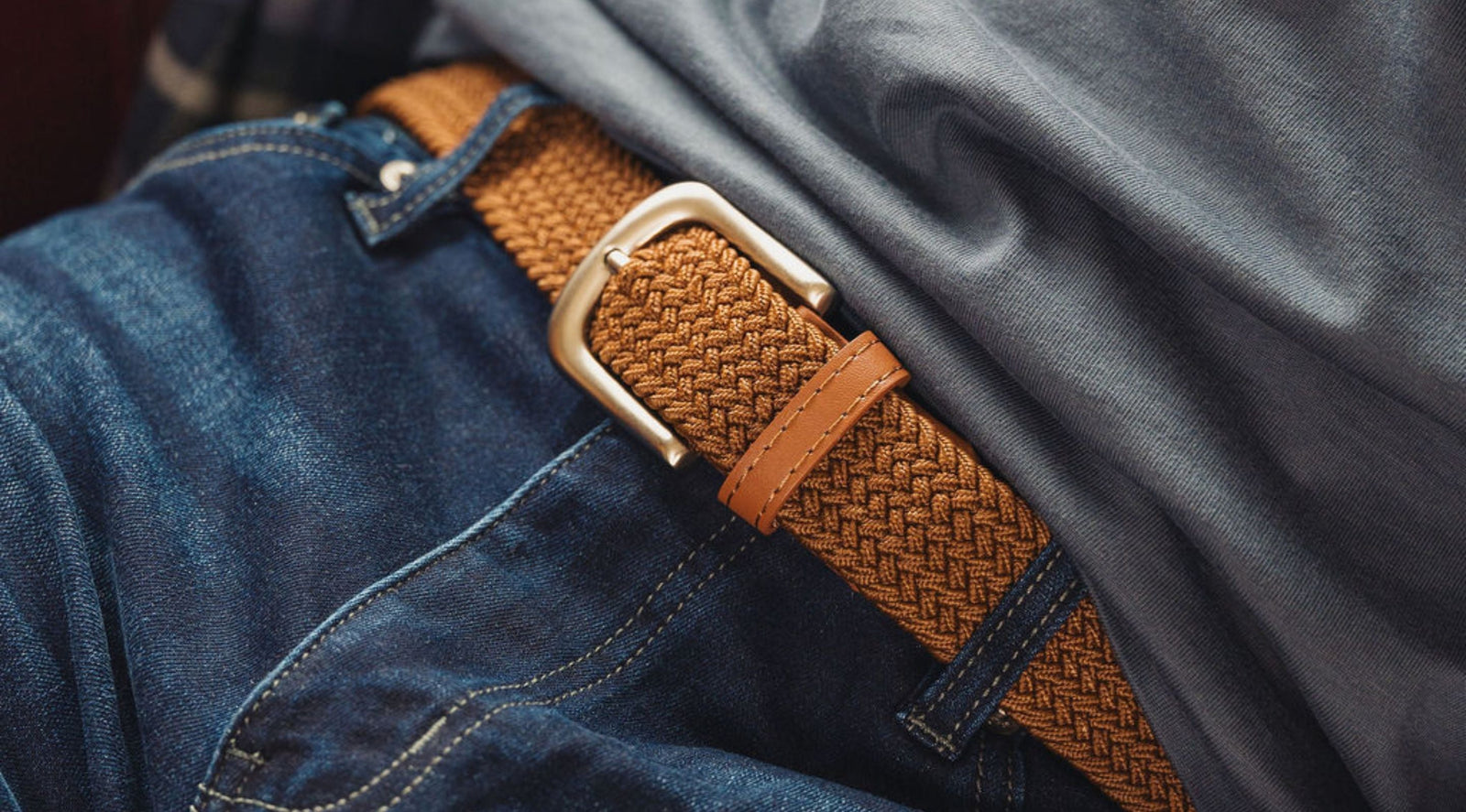 Braided tan belt with silver buckle threaded through blue jeans, gray shirt visible.