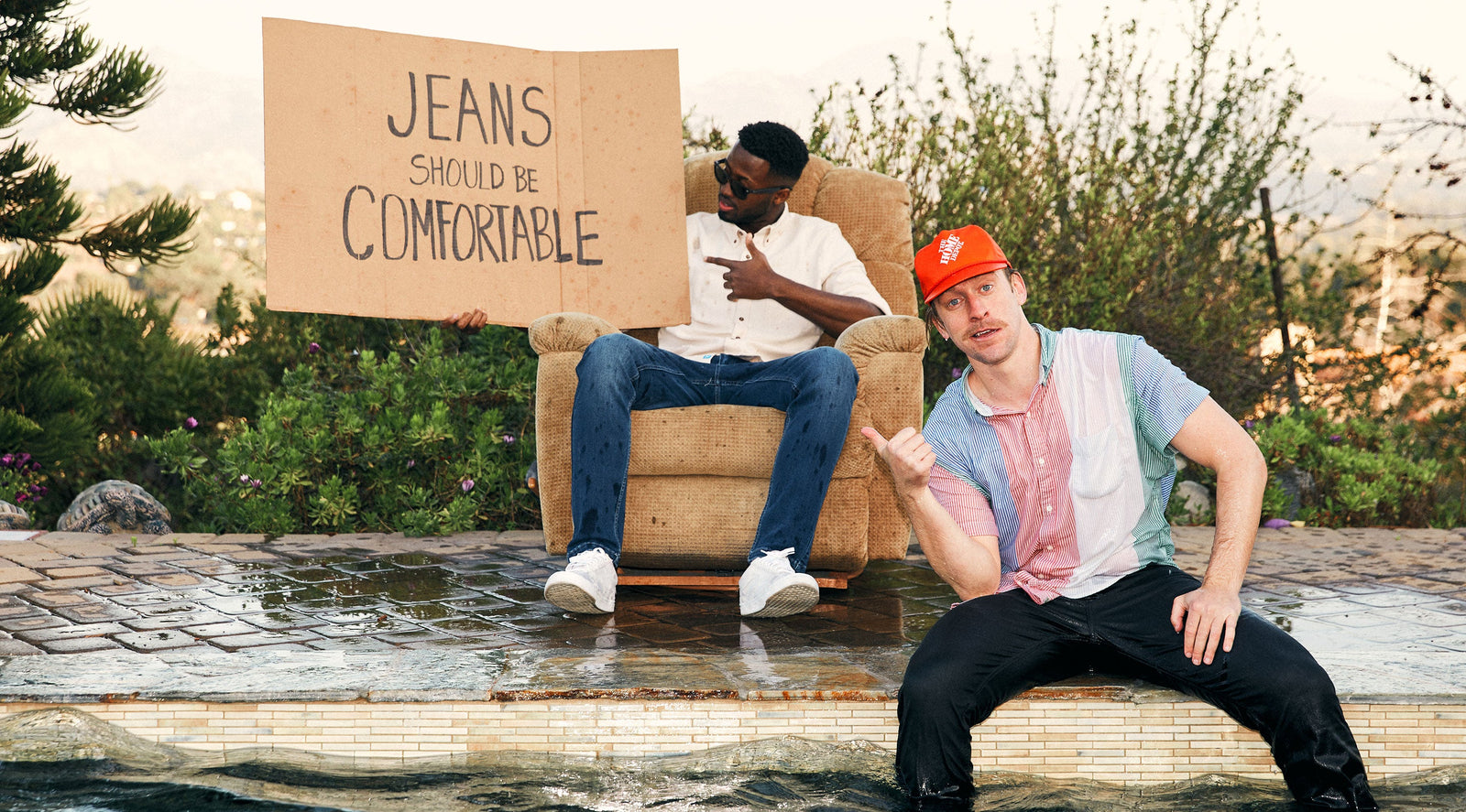 Two men by a pool; one sitting in an armchair holding a cardboard sign that reads 'JEANS SHOULD BE COMFORTABLE', the other sitting on the pool edge wearing a red cap and colorful shirt.