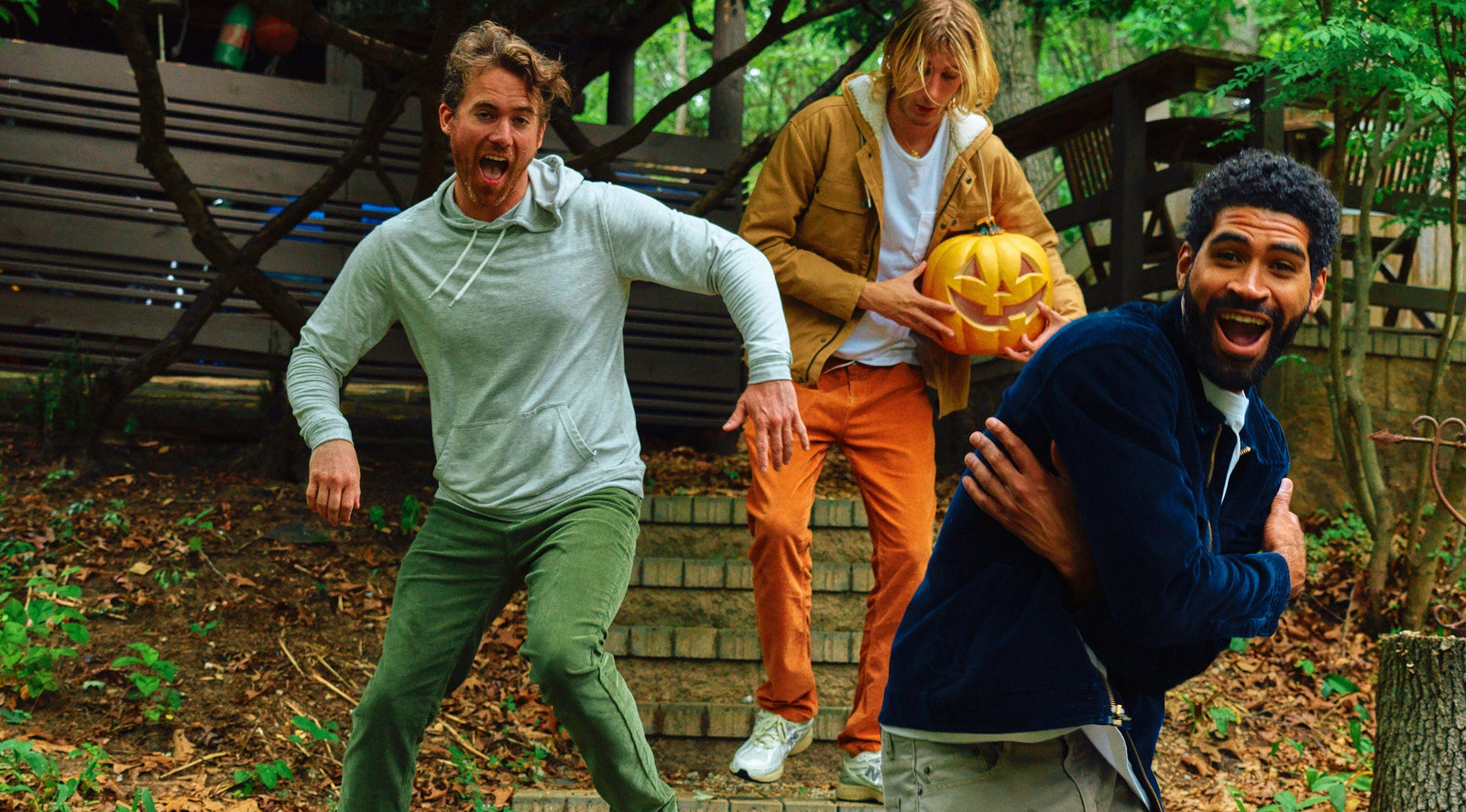 Three men outdoors on steps, one holding a carved Halloween pumpkin, all appearing joyful and playful.