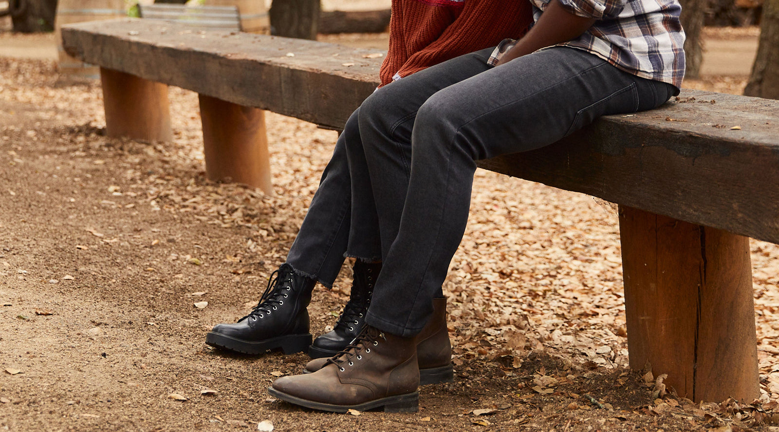 Two people sitting on a wooden bench outdoors wearing black denim pants and boots, with fallen leaves on the ground.