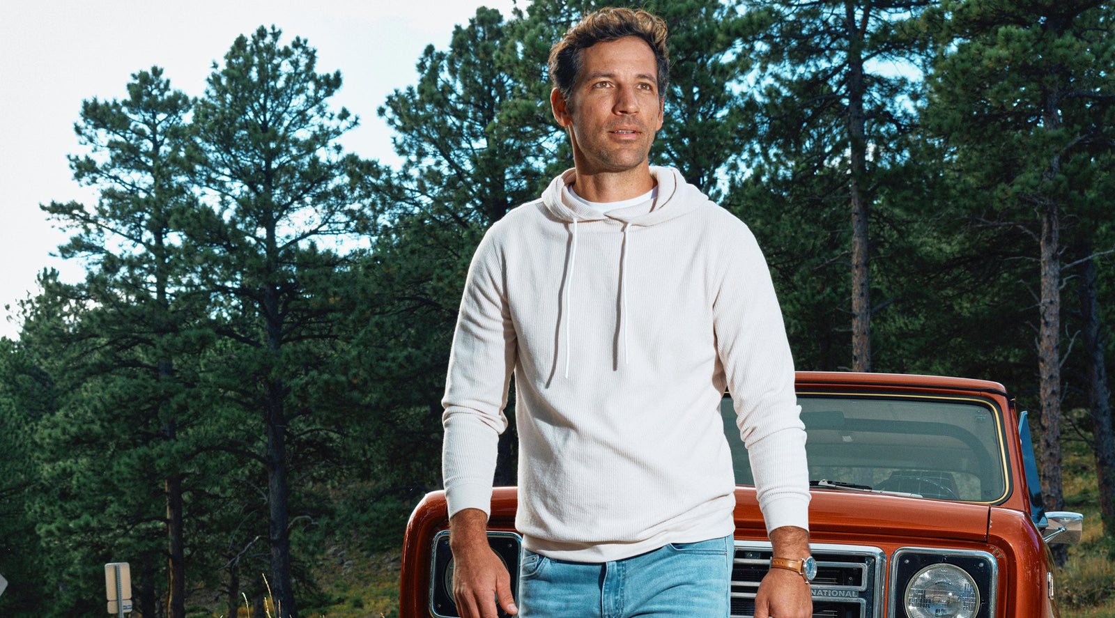 Man wearing a white hoodie standing in front of a red vintage truck with pine trees in the background