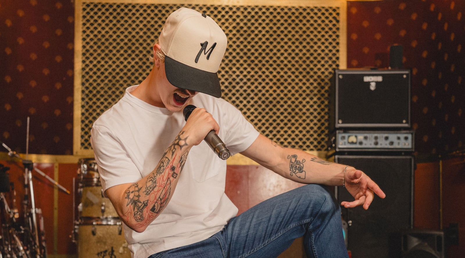 Front three-quarter view of cream-and-black 'M' cap showcasing embroidered M logo and black brim with rehearsal room backdrop