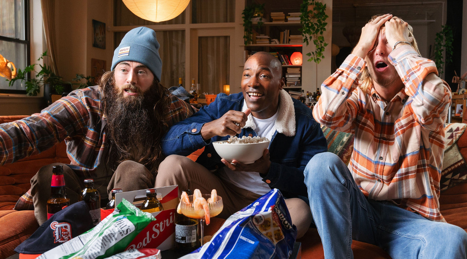Three men sitting on a couch with snacks and drinks, one wearing a blue beanie and plaid shirt, another in a denim jacket eating popcorn, and the third in a plaid shirt holding his head in surprise.