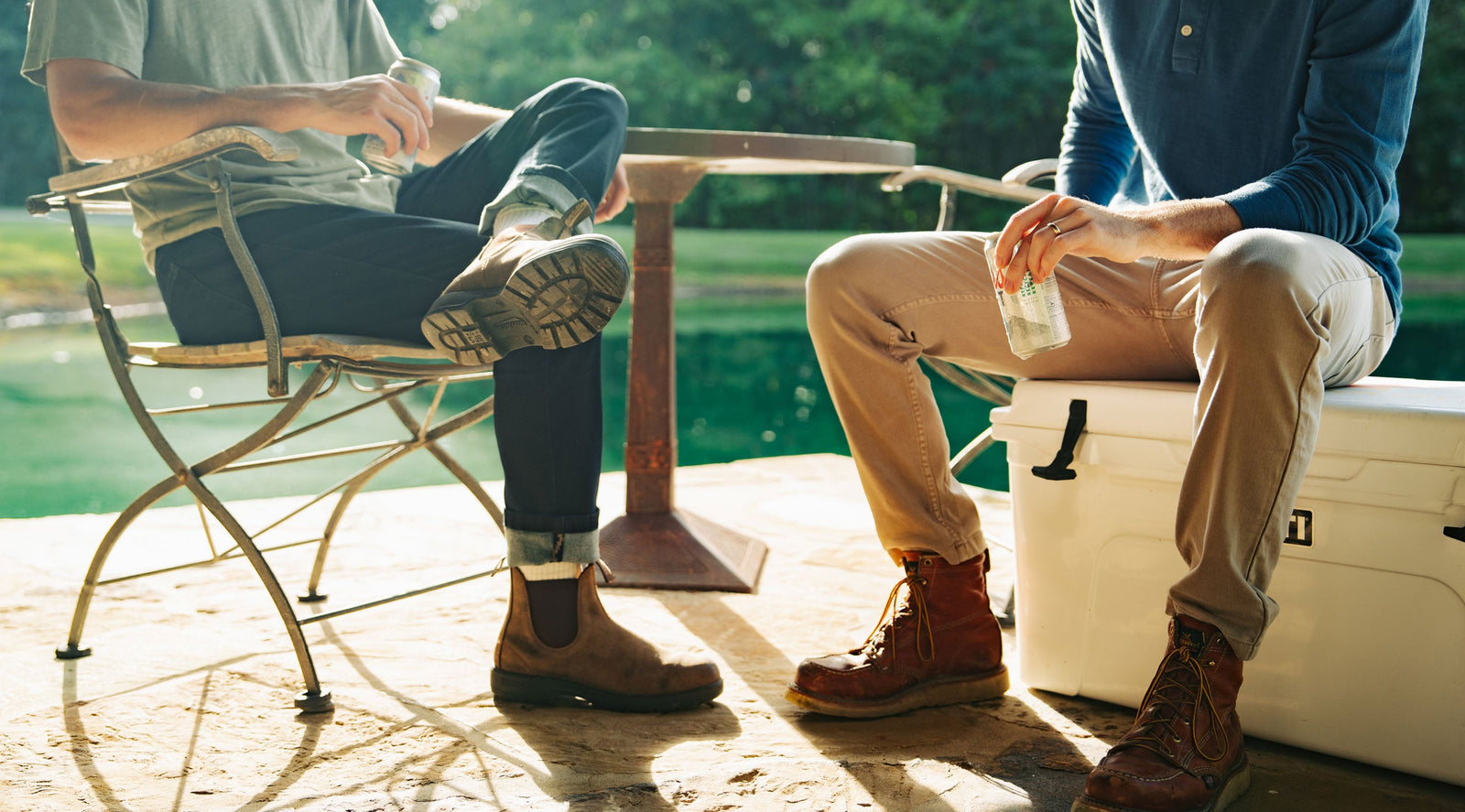 Two people sitting outdoors by a lake, wearing boots and holding canned drinks, one seated on a metal chair and the other on a cooler.