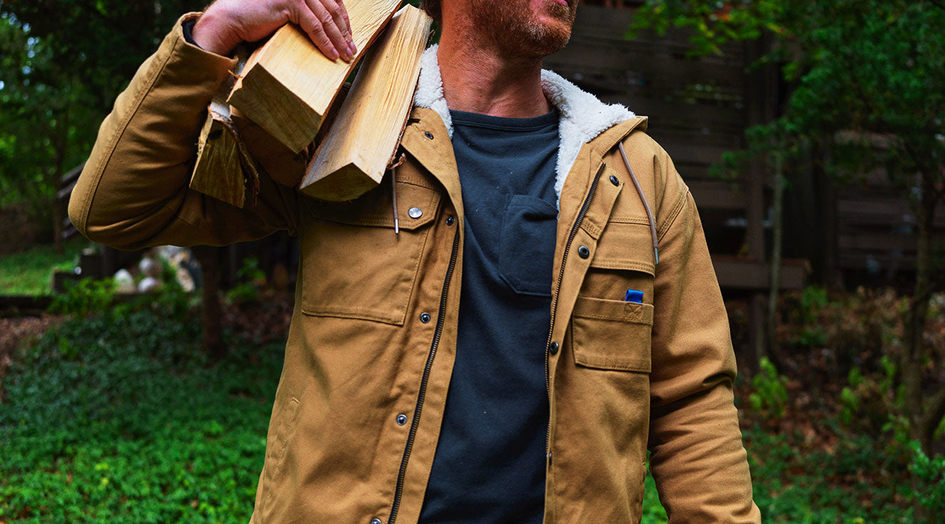 Man carrying split firewood over shoulder wearing tan sherpa-lined canvas jacket, close-up on torso and chest pocket.