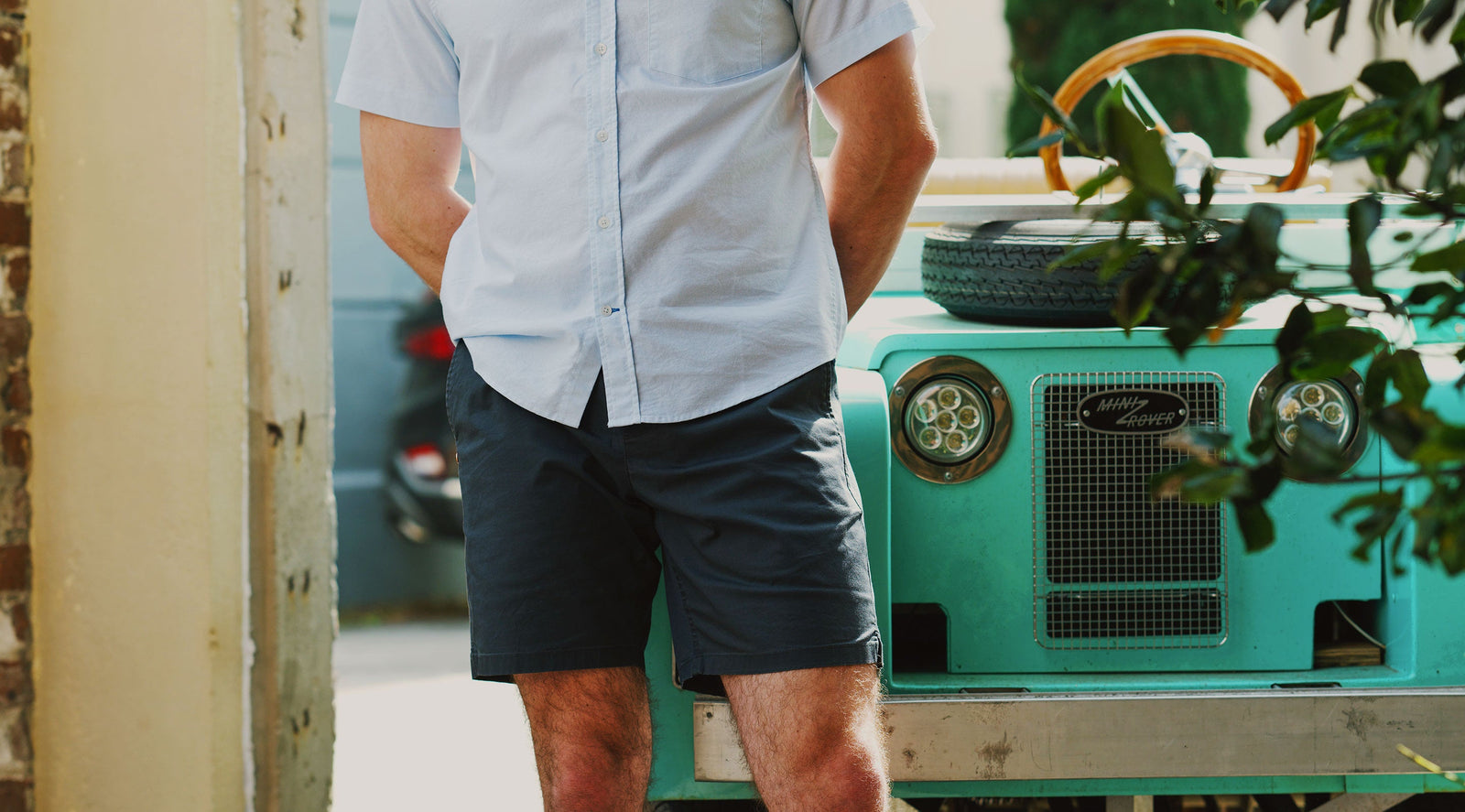 Man wearing navy shorts and a light blue shirt standing next to a turquoise vintage car with a spare tire on the hood.
