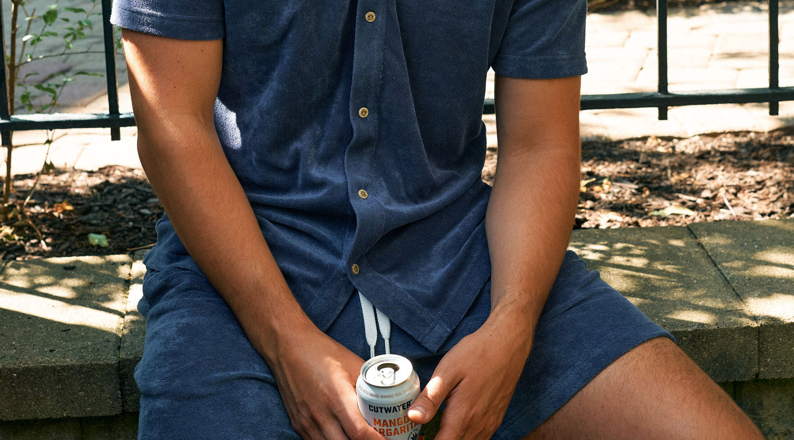 Person wearing a blue terry knit button-up shirt and matching shorts sitting outdoors on a stone ledge, holding a can labeled 'CUTWATER MANGO MARGARITA'.