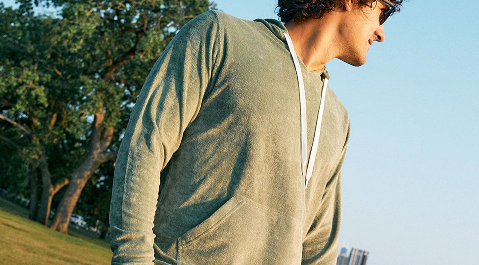 Man wearing a green terry knit hoodie with white drawstrings outdoors in a park with trees and buildings in the background