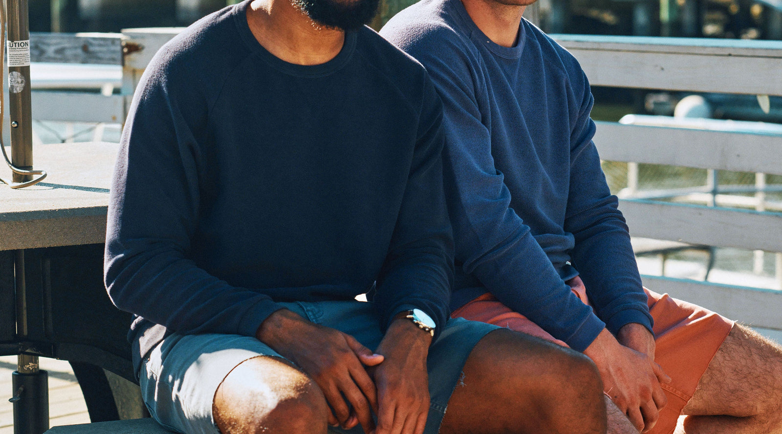 Two men sitting outdoors on a bench wearing navy and blue crewneck sweatshirts with shorts, one wearing a watch.