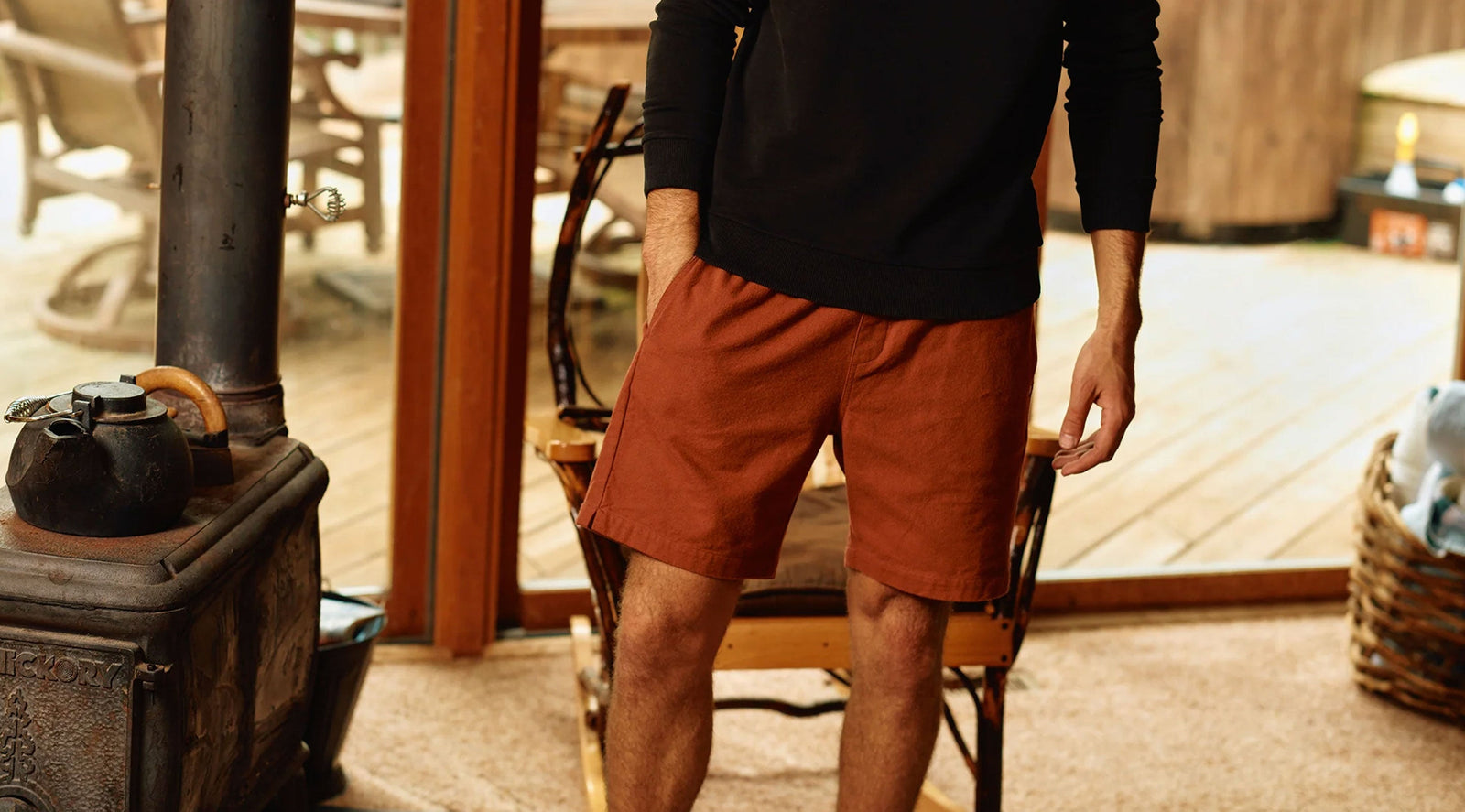 Front view of rust utility shorts with relaxed fit, mid-thigh length and side pockets, model standing indoors by wood stove