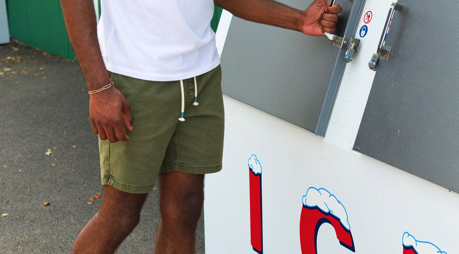 Person wearing green shorts and a white shirt opening a locked gray panel with two padlocks outdoors.