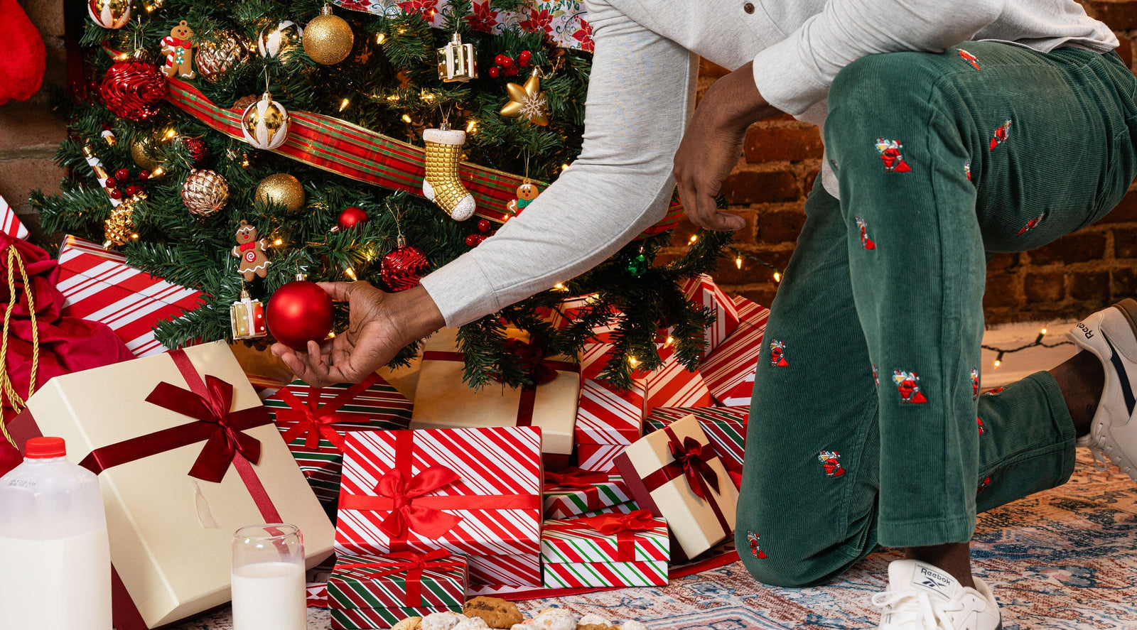 Person kneeling to hang a red ornament on a Christmas tree among wrapped gifts, milk and cookies; Reebok visible on shoe.