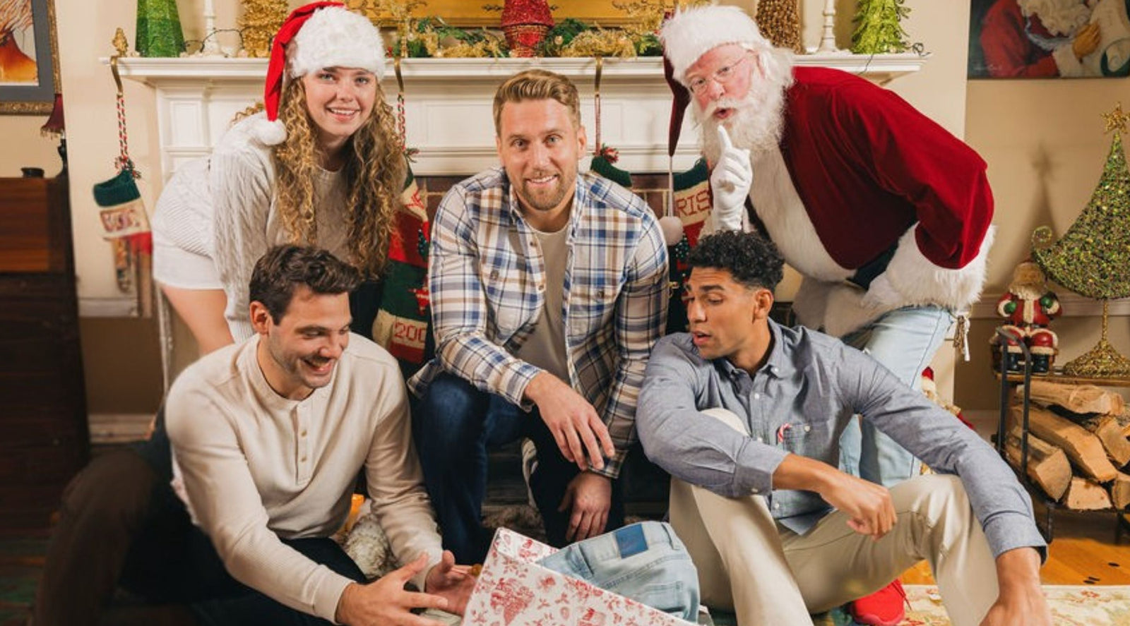 Five people, including someone dressed as Santa, opening a gift by a decorated mantel with stockings and holiday decor