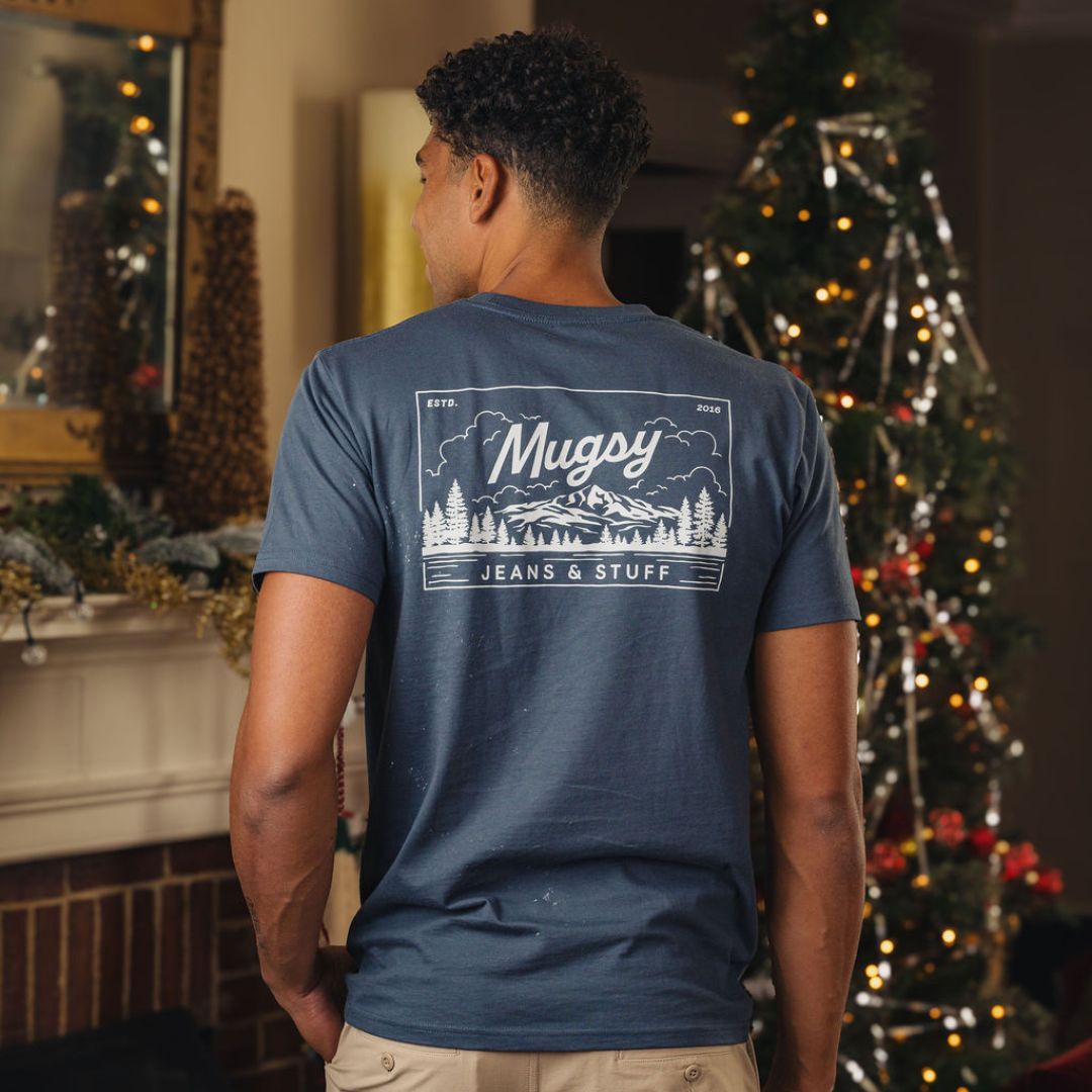 Man from back wearing navy T-shirt with white logo: Mugsy JEANS & STUFF ESTD. 2016, standing by decorated Christmas tree