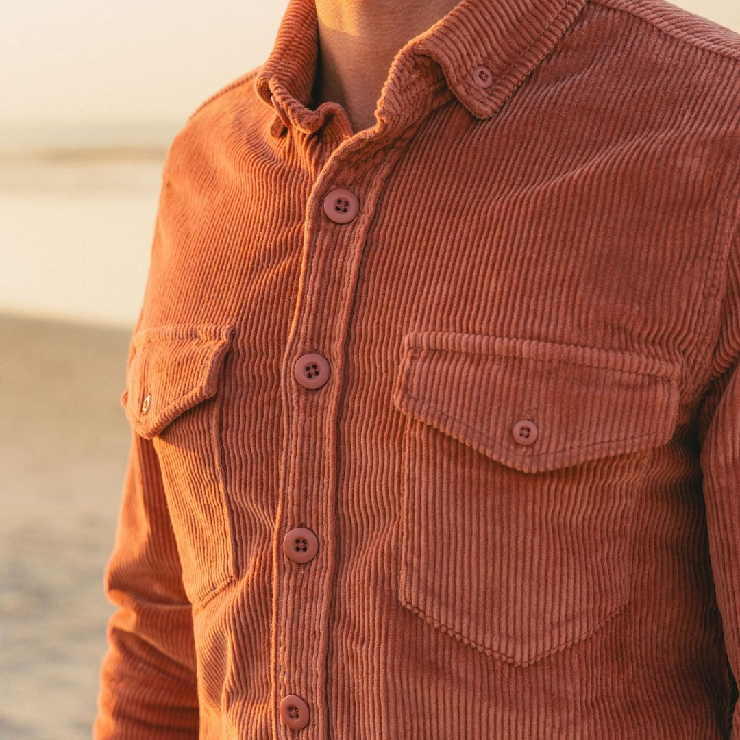 Rust-colored corduroy button-front shirt close-up, showing vertical ribbed texture, four buttons, and two flap chest pockets.