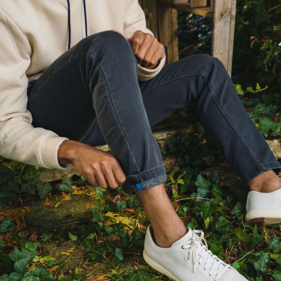 Seated person rolling up a jean cuff, wearing white sneakers and a cream hoodie amid ivy-covered garden steps.