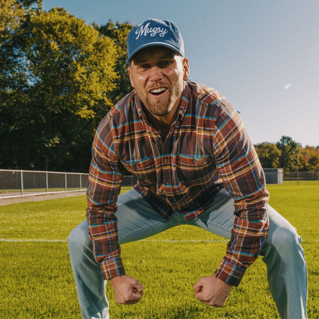 Athletic man in plaid shirt and Mugsy cap crouching on a sunlit grassy sports field, fists clenched, smiling.