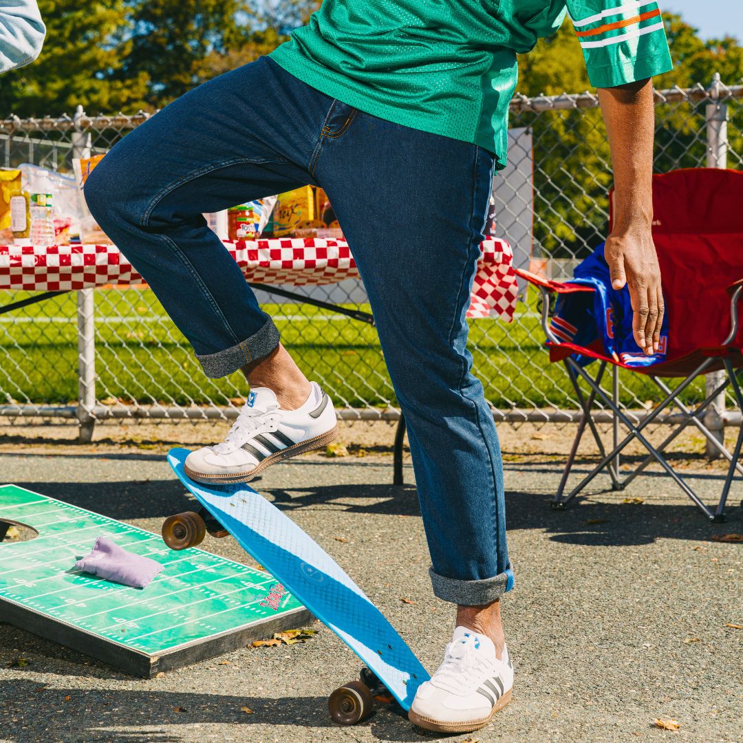 Person in rolled jeans and green jersey balancing on a blue skateboard beside a picnic table and folding chair