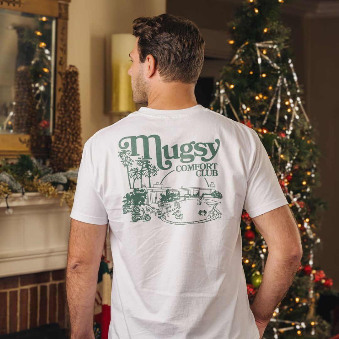 Back view of man wearing white T-shirt printed Mugsy COMFORT CLUB, standing by a decorated Christmas tree.