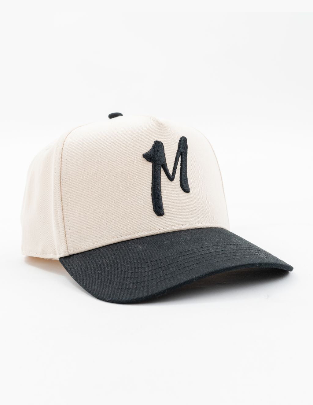 Cream baseball cap with black brim and embroidered M on front, angled on white background