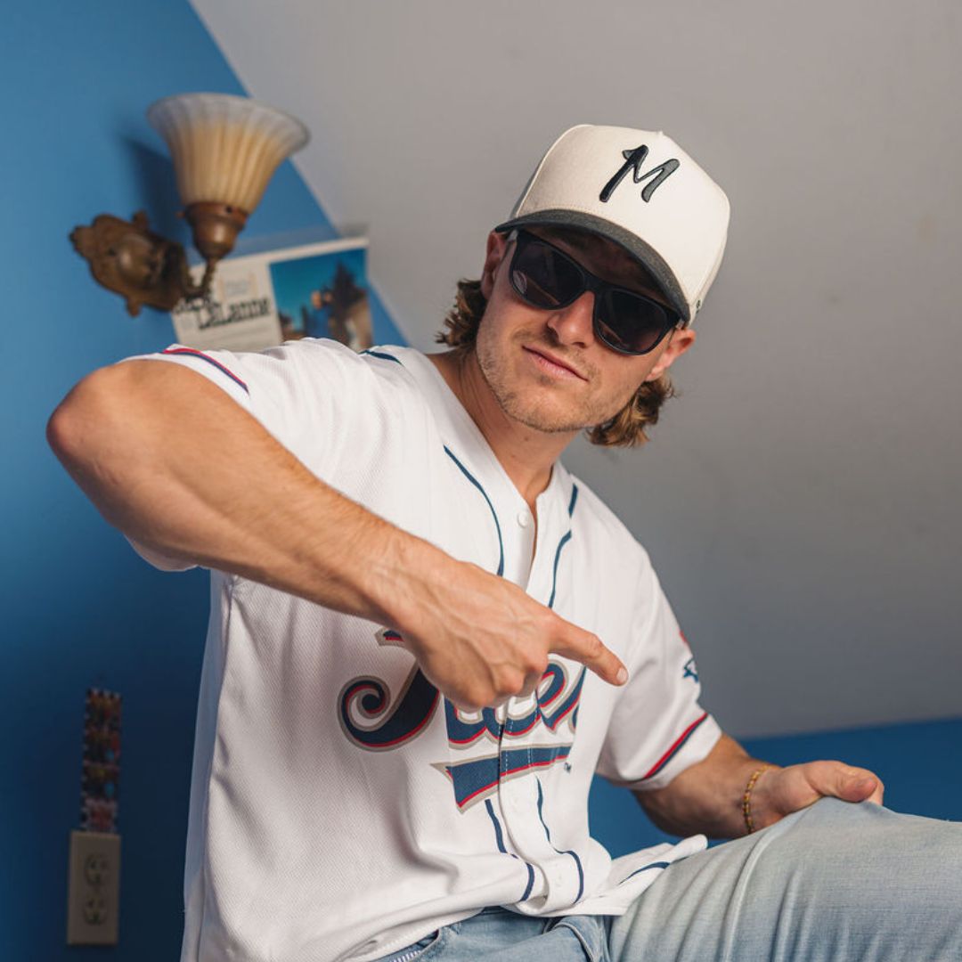 Young man in sunglasses and a baseball cap with M, pointing at his jersey while sitting in a blue-walled room.