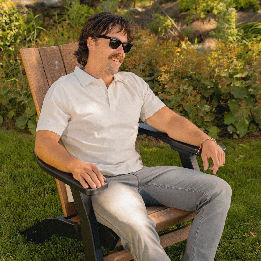 Relaxed man with mustache and sunglasses seated in a wooden Adirondack chair outdoors, wearing a light polo and jeans.