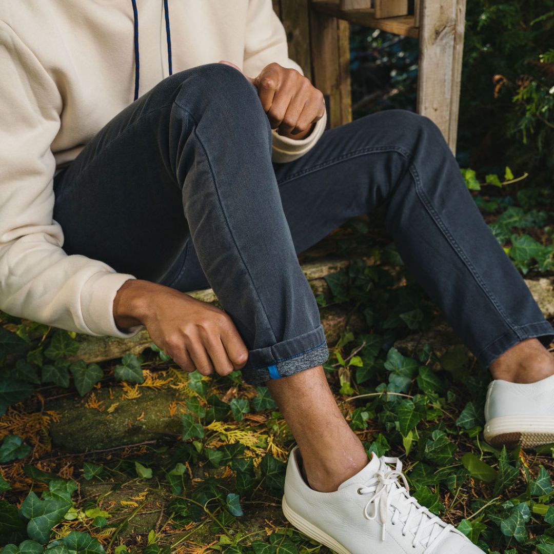 Person sitting on a wooden step outdoors, rolling up dark gray jeans; cream hoodie and white sneakers on ivy.