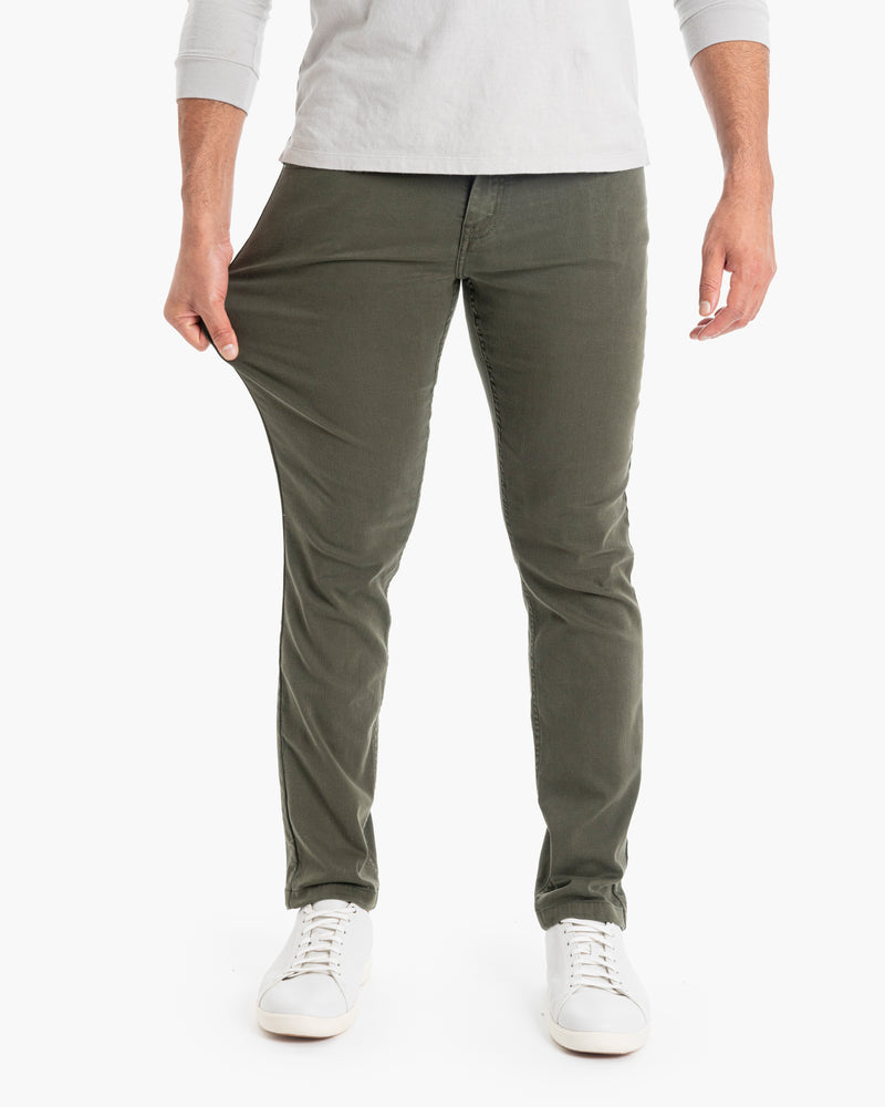 Men's olive-green stretch slim-fit pants shown on model pulling thigh to show fabric stretch, paired with white sneakers