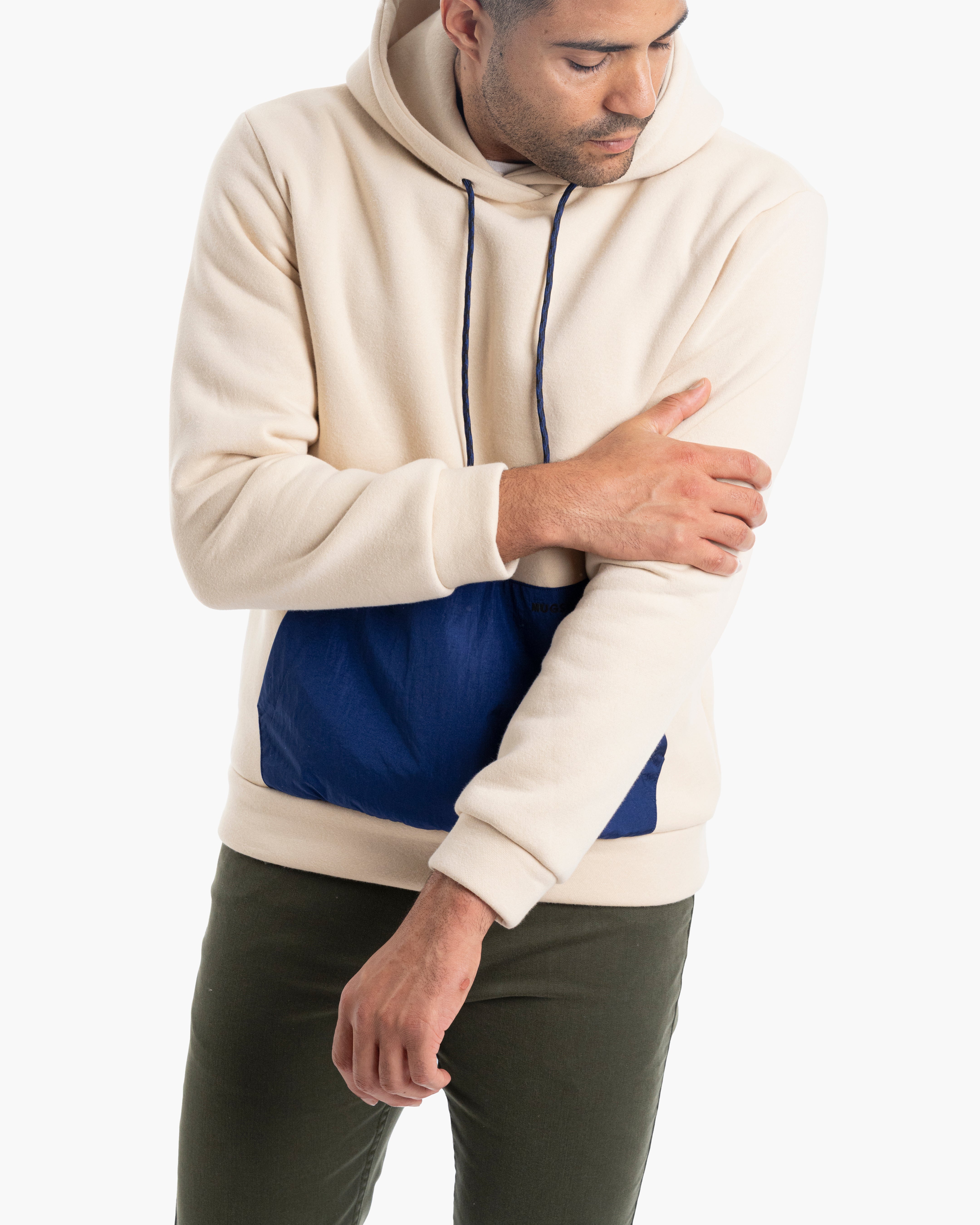 Man wearing beige hoodie with navy kangaroo pocket and navy drawstrings, adjusting his left sleeve while looking down.