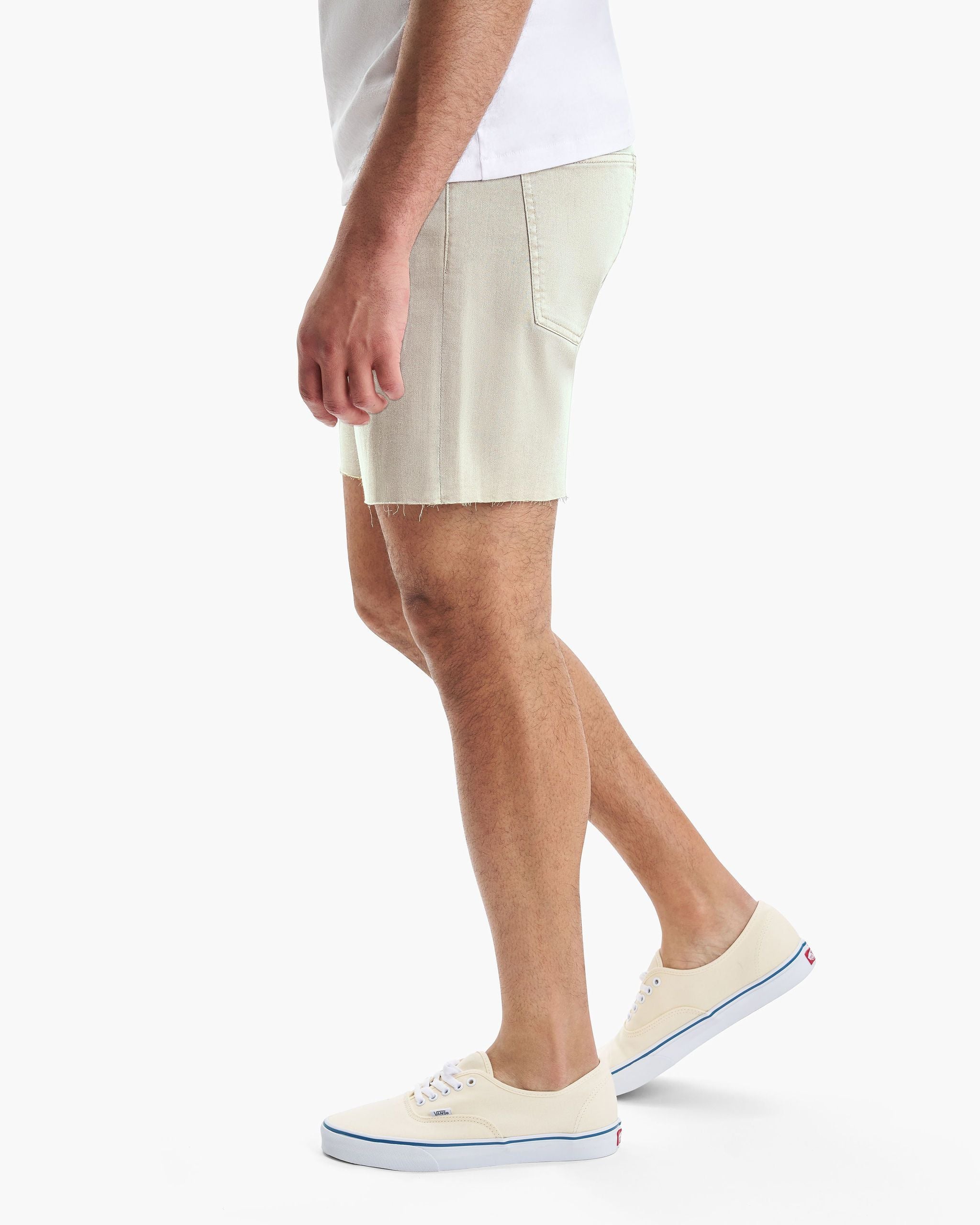 Side view of a person wearing beige denim shorts and cream-colored Vans sneakers with blue trim.