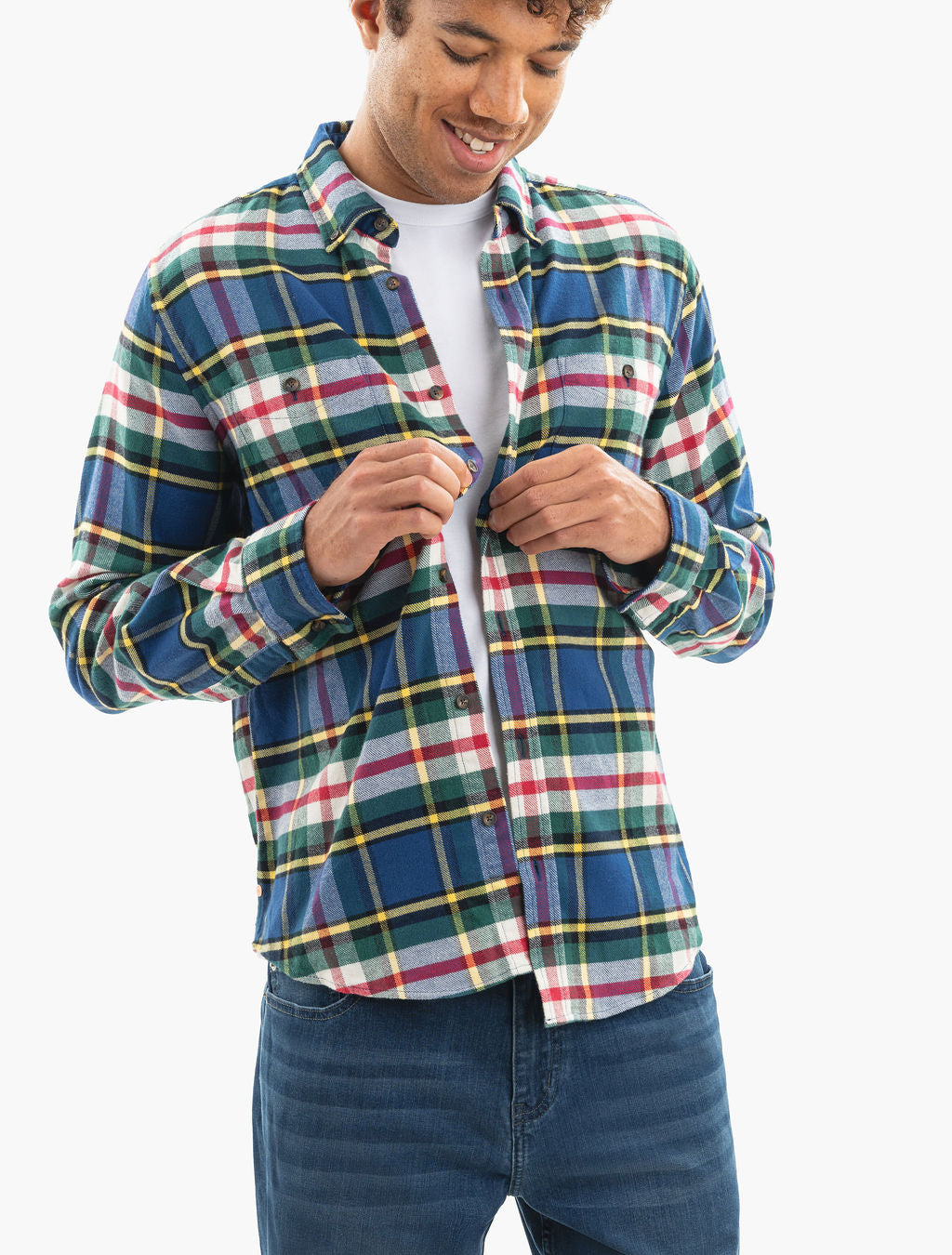 Young man buttoning a multicolor plaid flannel shirt over a white T-shirt, wearing blue jeans