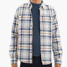 Smiling man shown from chin to hips wearing a navy T-shirt and beige-and-blue plaid button-up shirt, hands at sides.