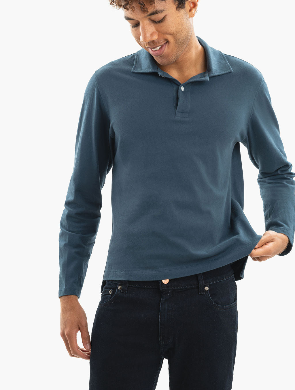 Male model wearing teal long-sleeve polo and dark jeans, pulling hem to show fit
