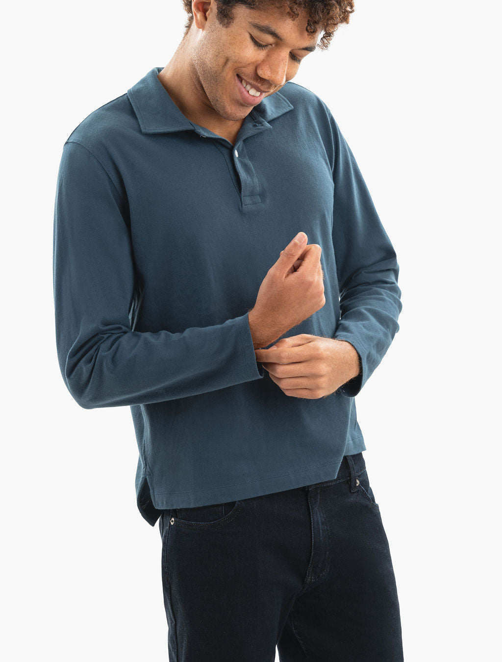 Smiling man adjusting the cuff of a teal long-sleeve polo shirt, shown from shoulders to hips.