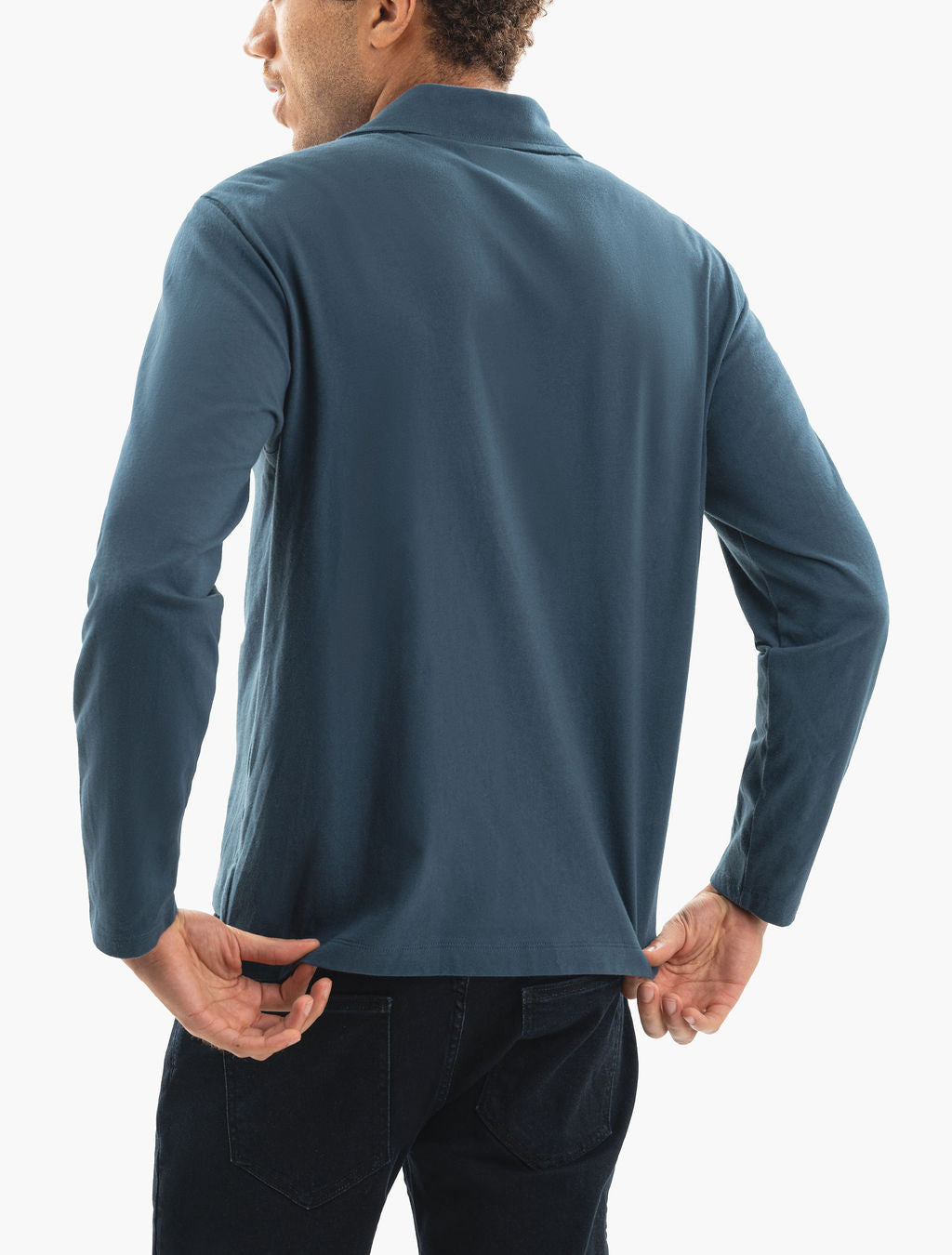 Man seen from the back wearing a teal long-sleeve polo shirt, pulling the hem to show the fit