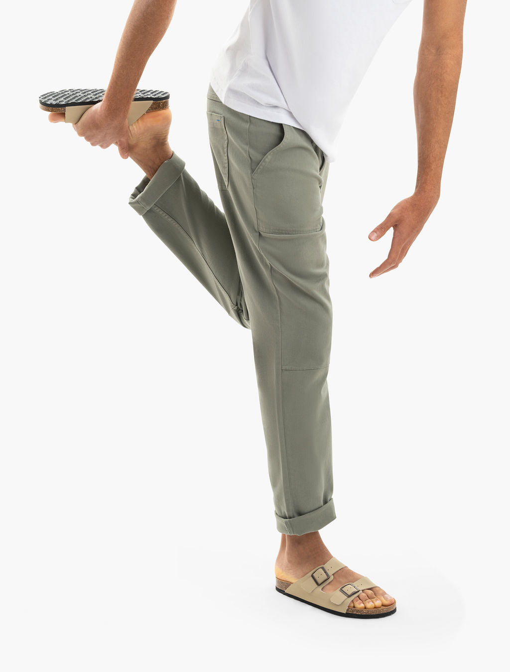 Person stretching, holding left foot behind; wearing cuffed olive-green pants and beige double-strap sandals.
