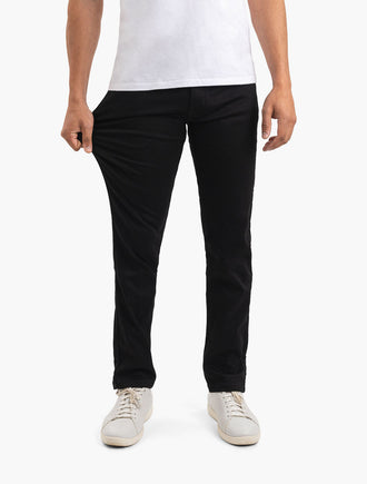 Men's black slim stretch pants shown waist-to-shoes; model pulls thigh to demonstrate fabric stretch.