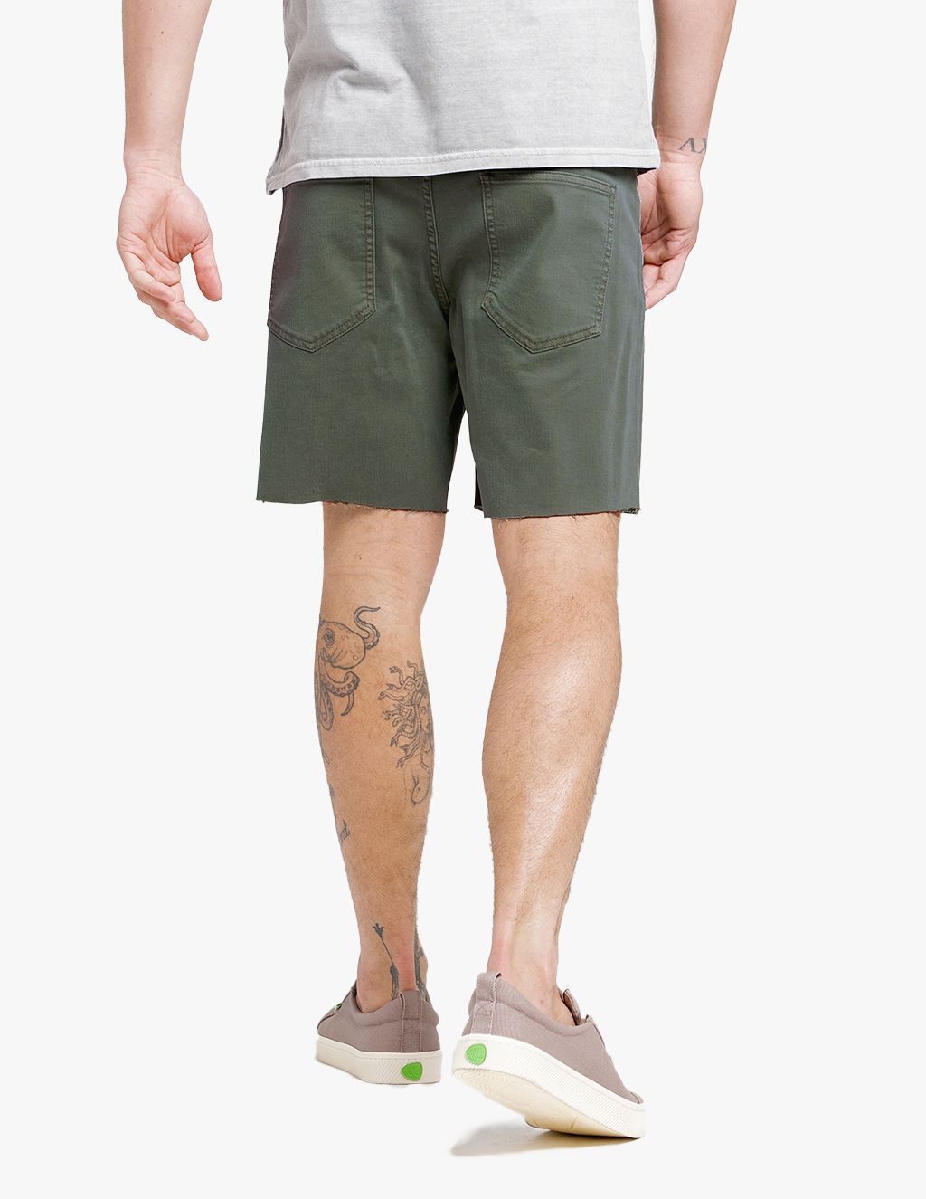 Rear view of a man wearing olive green shorts with back pockets, beige sneakers with green accents, and visible tattoos on his left calf and ankle.